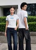 A man and a woman modeling matching white t-shirts with light blue trim, paired with dark jeans and white sneakers, standing outdoors in front of a modern building with green hedges.
